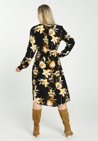 Robe noire à fleurs avec des fleurs jaunes et blanches, manches longues, taille ceinturée, longueur mi-mollet, associée à des bottes beiges jusqu'aux genoux.