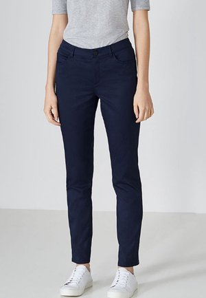 Frau trägt marineblaue, schmal geschnittene Hose, graues Kurzarmoberteil und weiße Sneaker, steht vor einem schlichten grauen Hintergrund.