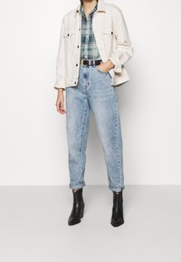 Veste en jean beige clair, chemise à carreaux vert et bleu, jean taille haute bleu clair, bottines noires. Design simple avec manches retroussées.