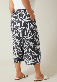 Geprinte cropped broeken met een donkere achtergrond en witte bloemenpatronen, elastische tailleband en zijzakken. Witte sandalen zichtbaar.