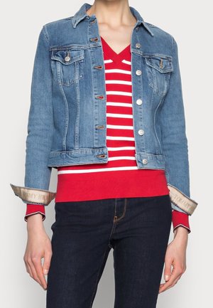 Persona che indossa una giacca di jeans con bottoni sopra un maglione a V a righe rosse e bianche, abbinato a jeans blu scuro.