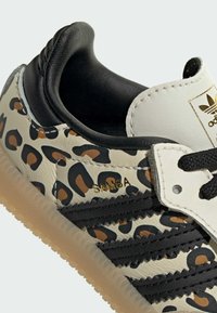 Zapatilla Samba con parte superior de cuero beige con estampado de leopardo, detalles en cuero negro, tres franjas negras y suela de goma color canela.