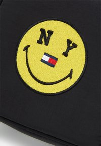 Un tessuto nero presenta un grande sorriso ricamato giallo con "NY" in nero sopra e un piccolo dettaglio con una bandiera rossa e blu sotto.