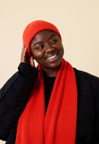 Red ribbed knit beanie and scarf, worn over a black quilted jacket, featuring a smooth texture and bold color, against a light background.