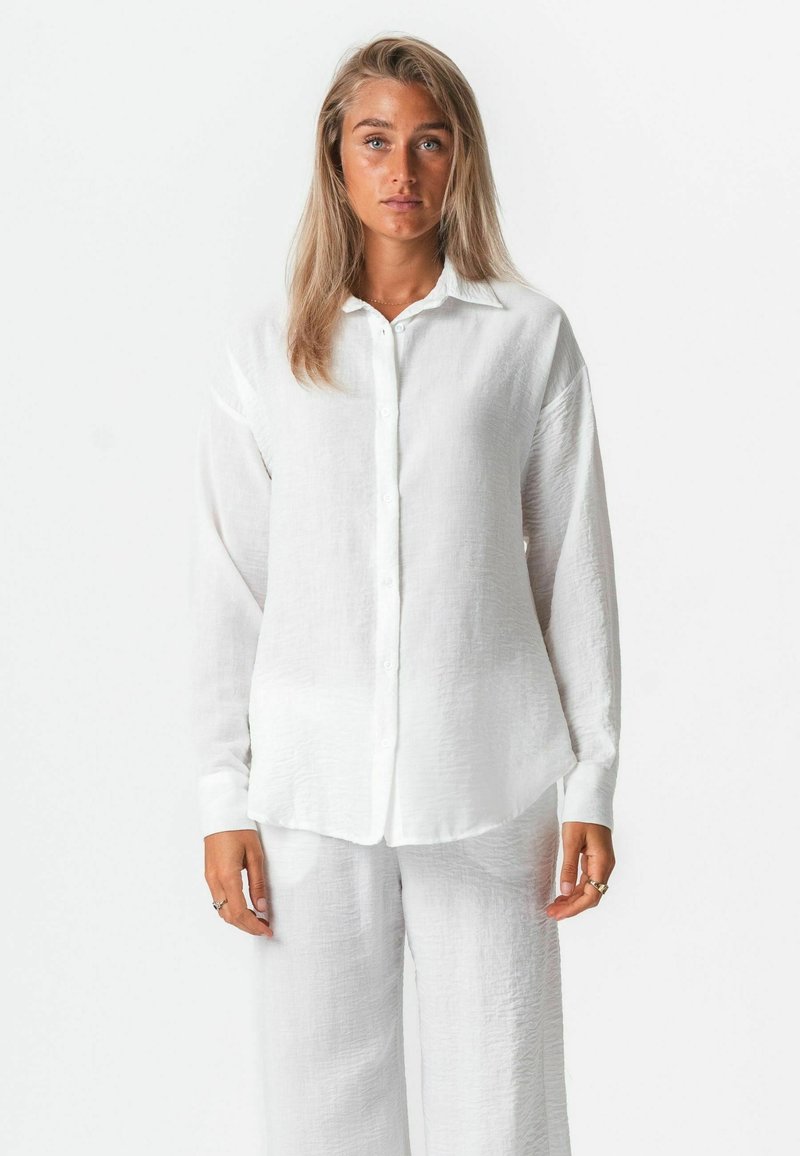 Jeune femme aux cheveux châtain clair portant une chemise blanche ample boutonnée et un pantalon blanc à jambes larges, debout devant un fond blanc uni.