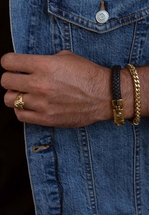 Zwarte gevlochten leren armband met gouden sluiting, gouden schakelarband en een gouden leeuwenkopring gedragen op een hand tegen een denim achtergrond.