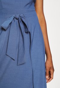 Primer plano de un vestido azul con un cinturón de tela atado a la cintura y el brazo derecho de una persona relajado a su lado.