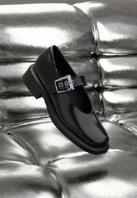 Black leather Mary Jane shoe with a square toe, a silver buckle accent, and a chunky low heel, set against a glossy silver background.