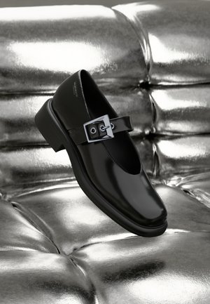 Black leather Mary Jane shoe with a square toe, a silver buckle accent, and a chunky low heel, set against a glossy silver background.