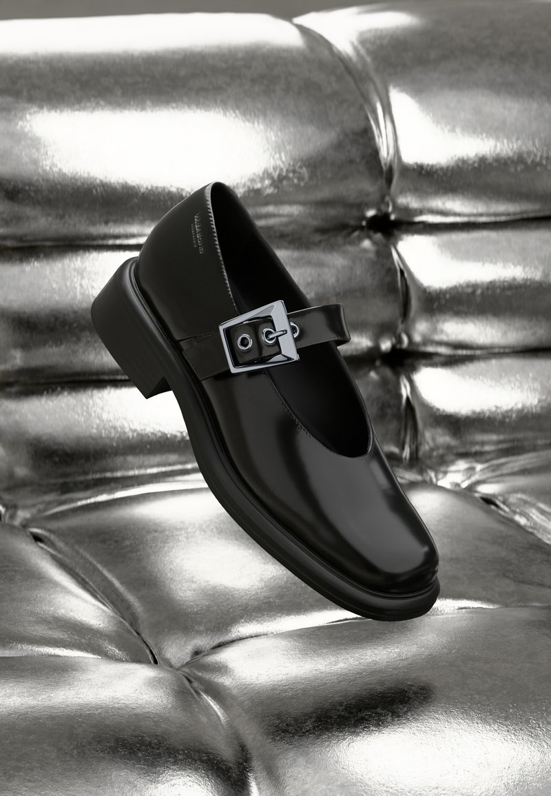 Black leather Mary Jane shoe with a square toe, a silver buckle accent, and a chunky low heel, set against a glossy silver background.