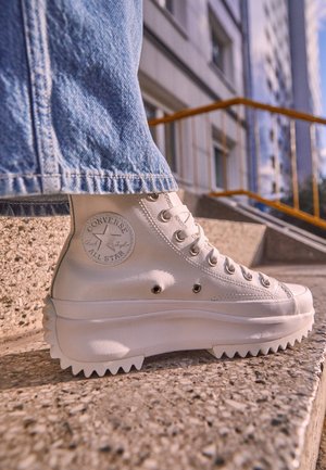 Zapatilla Converse blanca de caña alta con suela gruesa y acanalada, usada con vaqueros azules, sobre unos escalones de hormigón texturizado al aire libre.