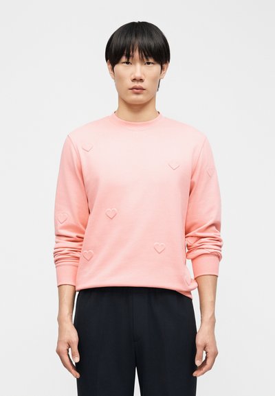 Νεαρό άτομο με ίσια μαύρα μαλλιά που φοράει ροζ φούτερ με ανάγλυφες καρδιές και μαύρο παντελόνι, στέκεται προς τα εμπρός.