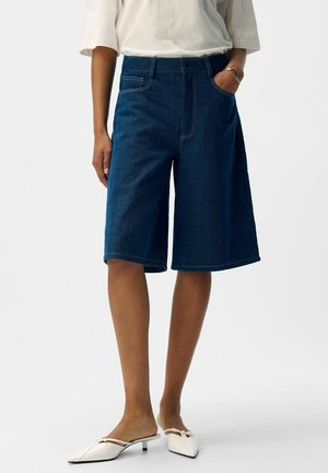 Personne portant un short en denim bleu foncé jusqu'au genou, un haut blanc, un bracelet argenté et des chaussures blanches à petit talon pointu avec un détail en forme de nœud.