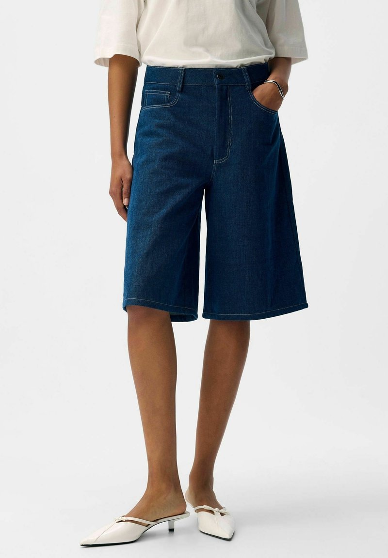 Personne portant un short en denim bleu foncé jusqu'au genou, un haut blanc, un bracelet argenté et des chaussures blanches à petit talon pointu avec un détail en forme de nœud.