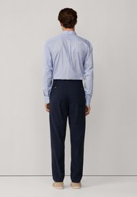 Uomo che indossa una camicia a righe azzurro chiaro infilata dentro pantaloni navy scuri, in piedi di spalle contro uno sfondo neutro.