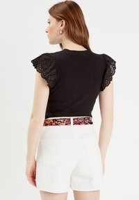 Top nero a coste con maniche in pizzo a ruffle, abbinato a pantaloni shorts bianchi. Cintura con motivo floreale visibile in vita. Design pulito, accenti minimi.