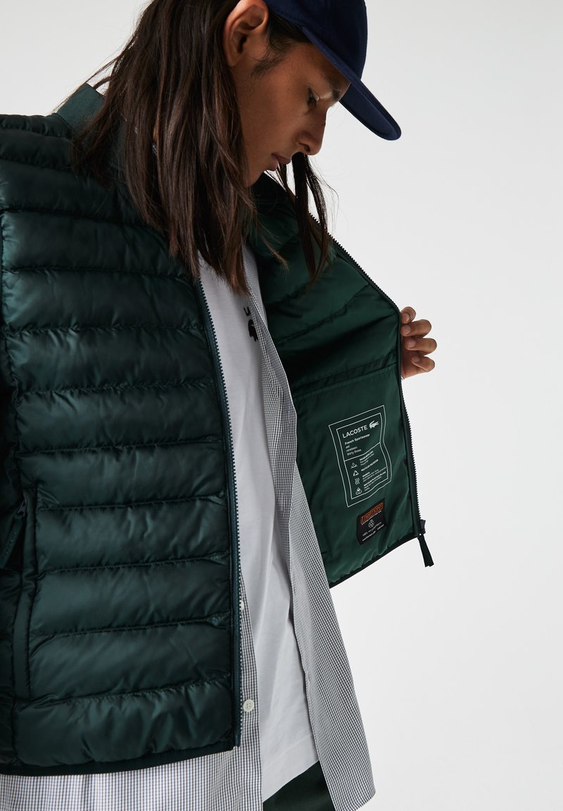 Chaqueta verde acolchada con un exterior brillante, cierre de cremallera y etiqueta interior visible. Lleva puesta sobre una camisa de rayas, que presenta un cuello alzado.