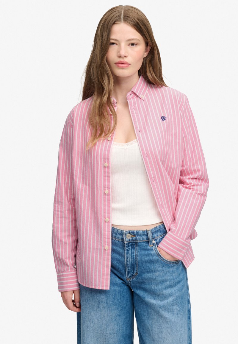 Giovane donna con capelli lunghi che indossa una camicia a righe rosa e bianca con bottoni sopra una canotta bianca e jeans blu, una mano in tasca.