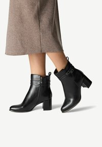 Black leather ankle boots with a high block heel, zip detail, and decorative straps. Smooth texture and fitted design.