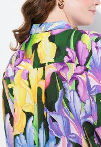 Una blusa floreale con un design vivace che presenta iris gialli, viola e rosa su uno sfondo verde, con colletto classico e vestibilità rilassata.
