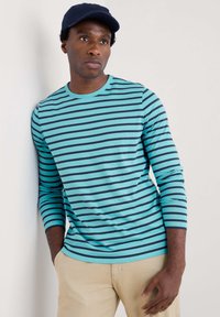 Tricou cu manșon lung, cu dungi în nuanțe de turquoise și bleumarin, din material de bumbac, cu guler rotund, textură moale, purtat cu pantaloni bej deschis.
