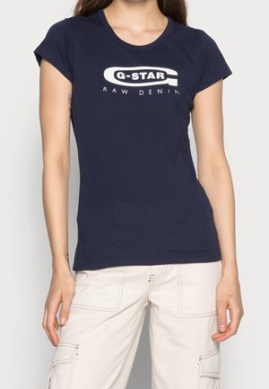 Kobieta w granatowej koszulce G-Star Raw Denim i jasnobeżowych spodniach typu cargo, stojąca z rękami swobodnie opuszczonymi wzdłuż ciała.