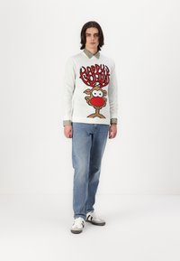 Suéter blanco de punto que presenta un reno de dibujos animados con una nariz roja y el texto "Felices Fiestas", combinado con jeans azul claro y zapatos a rayas negras.