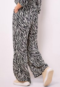 Persona che indossa pantaloni a zebrato bianco e nero a gamba larga con tasche e sneakers beige, in piedi contro uno sfondo chiaro.