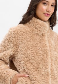 Beżowy płaszcz z sztucznego futra o fakturowanej, puszystej powierzchni, z wysokim kołnierzem i kieszeniami z przodu. Miękki materiał o ciepłym wyglądzie.