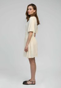 Robes beige avec des manches courtes et volantées, taille froncée et jupe fluide. Portée avec des sandales à lanières noires sur un mannequin devant un fond neutre.