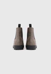 Een paar taupe suède Chelsea-boots met zwarte elastische zijpanelen en zwarte daadwerkelijk, weergegeven vanaf de achterkant op een witte achtergrond.