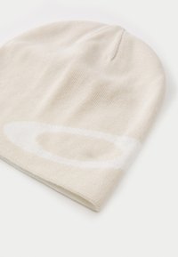 Creme strikket beanie-hue med en glat tekstur, der har et hvidt rundt logo-design. Stoffet er let og fleksibelt.