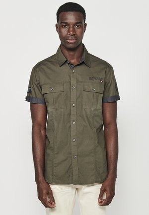 Camisa de algodón de manga corta en color verde oliva, con bolsillos frontales con botones, cuello oscuro en contraste y detalle de marca bordado.