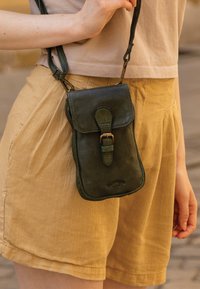 Green leather crossbody bag with a flap and buckle closure, featuring a zipper. The bag is worn with light yellow shorts.