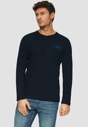 Marineblauwe henley t-shirt met lange mouwen in zachte stof, met een drukknoopsluiting van drie knopen, voorzien van een logo aan de voorkant en een comfortabele pasvorm. Gecombineerd met lichtblauwe jeans.