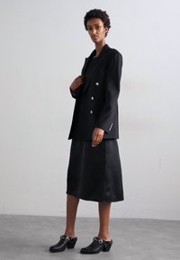 Zwarte dubbelgeknoopte blazer met zilveren knopen, gecombineerd met een zwarte rok tot de knie en zwarte hoge klompen met ronde accenten.