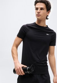 Νεαρός άνδρας με μαύρο αθλητικό μπλουζάκι και σορτς που κρατά ένα μαύρο παγούρι νερού, κοιτώντας ελαφρώς προς το πλάι μπροστά σε απλό φόντο.