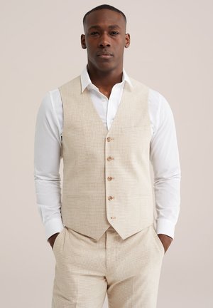 Mann trägt beigefarbenes, kariertes Weste und passende Hose über einem weißen Hemd, steht mit Händen in den Taschen.