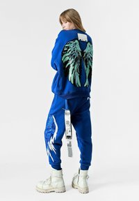 Sudadera azul con gráficos de alas verdes y negras en la parte posterior, combinada con pantalones de chándal azul a juego que presentan un diseño plateado y etiquetas. Botas crema.