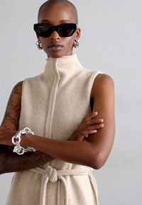 Abito senza maniche beige in tessuto testurizzato, con colletto alto, chiusura frontale con zip e vita legata. Il modello indossa occhiali da sole neri audaci e orecchini d'argento.