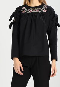 Svart blus med blommig broderi vid halsringningen, med axelband och långa ärmar, tillverkad av ett mjukt textil.