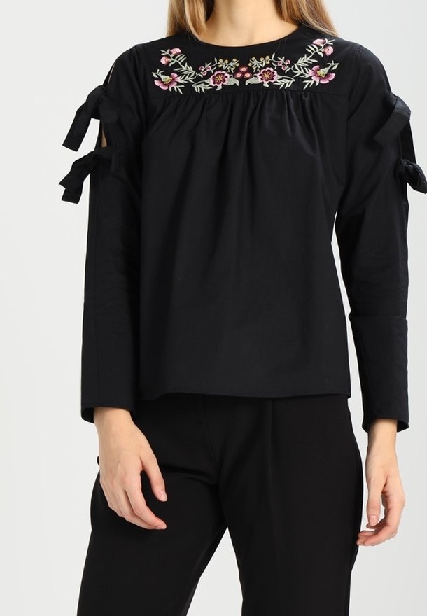 Svart blus med blommig broderi vid halsringningen, med axelband och långa ärmar, tillverkad av ett mjukt textil.
