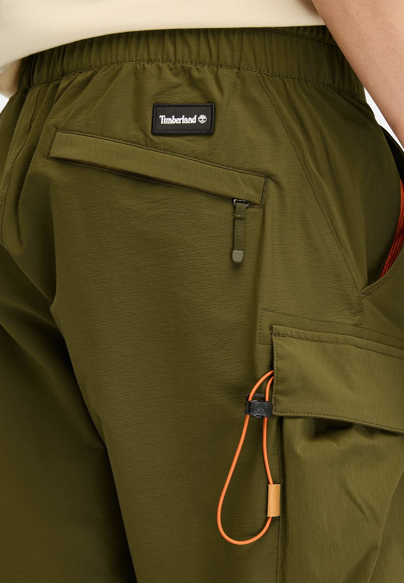Grüne Cargo-Hosen mit elastischem Bund, ausgestattet mit einem Timberland-Logo, Reißverschluss- und Klappentaschen sowie einem orangefarbenen Kordelzug-Detail.