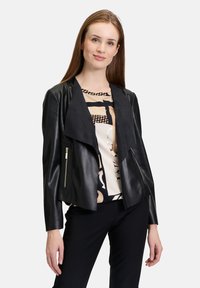 Veste en cuir noir avec un col drapé, des poches zippées et une texture lisse, assortie à un haut à motifs et à un pantalon noir élégant.