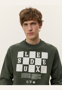 Sudadera verde con cuello redondo, que presenta un patrón de tablero de ajedrez blanco y letras impresas en negrita. Textura suave con un ajuste relajado.