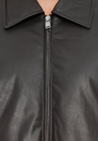 Veste en cuir noir avec une texture lisse, un large col et une fermeture éclair centrale en argent, avec des lignes épurées et sans motifs visibles.