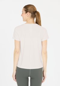Mujer con una coleta marrón que lleva una camiseta blanca de manga corta suelta y leggings gris oscuros, de pie y de espaldas, sonriendo de lado.