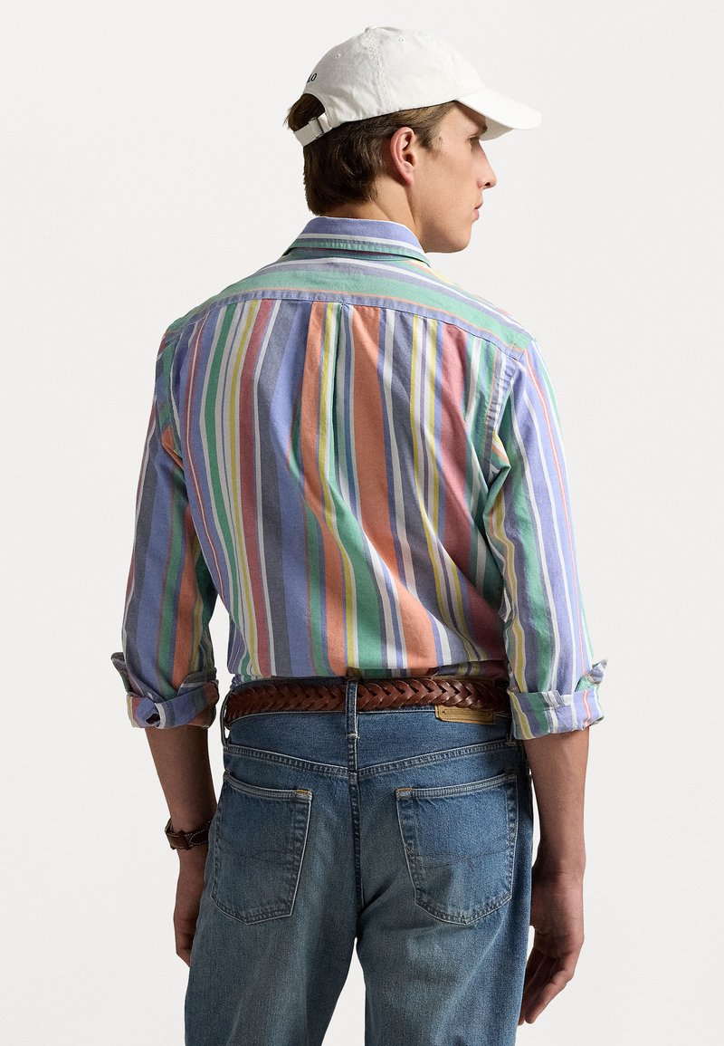 Langärmliges Hemd mit mehrfarbigen vertikalen Streifen in Pastellfarben, kombiniert mit Jeans und einer weißen Kappe. Es hat einen Button-Down-Kragen.