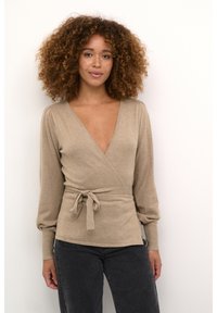Top envolvente beige en tejido de punto suave, con un escote en V profundo, mangas largas abullonadas y detalle de cintura atada. Combinado con jeans gris oscuro.