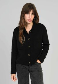 Cardigan noir à boutons fabriqué dans un matériau doux, avec un col, deux poches avant et de grands boutons marron. Manches longues avec détail au niveau des poignets.